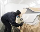 Pracownik w masce i kombinezonie maluje samochód w kabinie lakierniczej. Czas schnięcia bazy zależy od warunków.