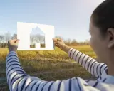 Szukasz idealnej działki pod dom 70m2? Kobieta trzyma wycięty z papieru dom na tle słonecznego pola i drzew.