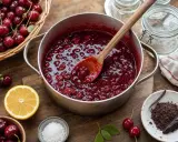 Une marmite déborde de confiture de cerises maison, prête à être mise en pot. Des cerises fraîches, du citron et du chocolat râpé complètent la scène.