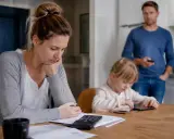Zmartwiona kobieta z kalkulatorem przy biurku, dziecko pochłonięte telefonem, mężczyzna stoi z tyłu. Czy należy się opieka nad dzieckiem gdy żona nie pracuje?