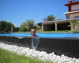 Niña sonriente en una piscina desbordante, con agua cayendo sobre piedras blancas y césped verde. Al fondo, una casa moderna y árboles frondosos bajo un cielo azul.
