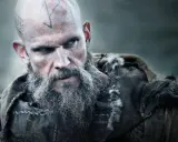 Uhtred z Bebbanburga, wojownik z tatuażami na głowie i długą, siwą brodą, spogląda groźnie.