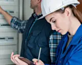 Kobieta w kasku i niebieskiej kurtce spisuje protokół odbioru instalacji elektrycznej, podczas gdy mężczyzna wskazuje na rozdzielnicę.