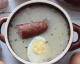 Aromaticzny zakwas na żurek w glinianym naczyniu, z kiełbasą i jajkiem. Obok czosnek, cebula i jajka.