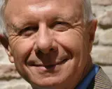 Primo piano sorridente di padre Alberto Maggi, con rughe d'espressione e capelli grigi, sullo sfondo di un muro di mattoni.