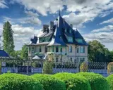 La villa Demoiselle, une demeure de caractère avec ses toits pointus et sa façade claire, se dresse fièrement derrière une grille ouvragée et des buissons taillés.