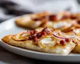 Tranches de flammekueche recette originale sur une assiette blanche, garnie de lardons, oignons rouges et fromage crémeux.