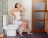 Ciężarna kobieta siedzi na toalecie, trzymając się za brzuch i czoło. W łazience widać bidet, co sugeruje lewatywa przed porodem.