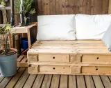 Meble z palet na balkon: wygodna sofa z szufladami, białe i szare poduszki, rośliny w doniczkach, drewniana ściana i podłoga.