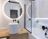 Elegante bagno senza finestra con lavabo bianco, specchio rotondo illuminato e doccia con finiture nere.