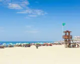 Sonniger Strand in Rota, Spanien, mit vielen bunten Sonnenschirmen und Menschen. Ein Rettungsschwimmerturm mit grüner Flagge steht am Meer.