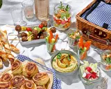 Leckere Picknick-Snacks: Fleischbällchen-Spieße, Gebäckstangen, Hummus, Gemüsesticks und süße Schnecken auf einer Decke.