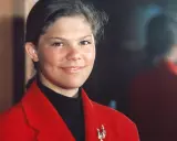 Kronprinsessan Victoria ung, med ett varmt leende, bär en röd kavaj och en svart polotröja. En vacker brosch pryder kavajen.