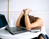 Personne épuisée, tête sur les bras, téléphone à la main, devant un ordinateur portable. L'effet écran de la fatigue est palpable.