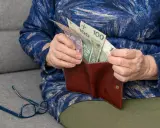 Dłoń starszej osoby wkłada pieniądze do portfela. Widać banknoty 100 zł. To wsparcie dla osób niepełnosprawnych, jak 500 plus dla niepełnosprawnych.