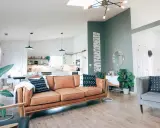 Jasny salon z brązową kanapą i szarym fotelem. Dominują kolory zimne, takie jak zieleń na ścianie i szarość mebli.