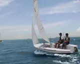 Deux jeunes hommes naviguent sur un dériveur 420 par une journée ensoleillée. D'autres voiliers sont visibles au loin.