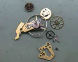 Parti di un orologio: ingranaggi, un bilanciere e altri componenti metallici disposti su una superficie grigia.