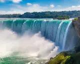 Potężny wodospad Niagara w całej okazałości. Woda spada kaskadami, tworząc mgłę. W tle drzewa i budynek.