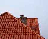 Ceglany komin wystaje ponad dach z czerwonej dachówki. Wlot do komina z pieca znajduje się na wysokości dachu.
