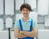 Un homme souriant en salopette bleue, entouré de chauffe-eau blancs, prêt à vous aider à choisir son chauffe eau.
