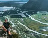 Man sitter på en klippa med en fantastisk utsikt över ett meandrande vattendrag och fjällandskap i Sarek. Aktse till skierfe!