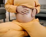 Schwangere Frau hält ihren Bauch, spürt den Druck im Unterleib. Gelber Jogginganzug, blaue Nägel.