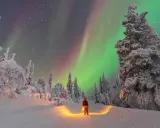 Un uomo con lanterna, vestito da Babbo Natale, ammira l'aurora boreale in Lapponia. Un paesaggio innevato e magico, perfetto per chi cerca dove andare in Lapponia.