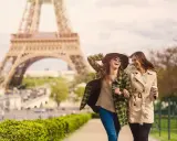 Due amiche ridono passeggiando a Parigi, con la Torre Eiffel sullo sfondo. Perfetto per capire cosa mettere in valigia per Parigi: cappotti leggeri, cappello e occhiali da sole.