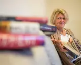 En kvinna med blont hår sitter med en bok i handen, omgiven av fler böcker. Hon ler mot kameran, som om hon delar en hemlighet från de fenomenala fruntimren på Grand Hotel.