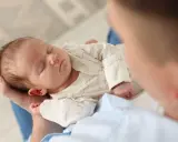 Un neonato dorme sereno tra le braccia di un genitore, cullato da suoni che ricordano i rumori bianchi, creando un'atmosfera di pace.