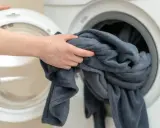 Ręka wkłada szary koc do pralki. Tak jak prac koce, by były czyste i pachnące.