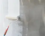 Rodillo blanco aplica pintura a pared gris. Se ve el proceso de cuantas manos de pintura se le da a una pared para un acabado perfecto.