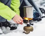 Tänd ett bensinkök i snön för att värma upp en kastrull. En person i en ljusgrön jacka och vandringskängor förbereder mat utomhus.
