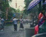 Ulewny deszcz na Bali podczas pory deszczowej. Mężczyźni w tradycyjnych strojach niosą drewniane kije, a widzowie chronią się pod kolorowymi parasolami.