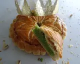 Galette des rois pistache dorée, une part coupée révèle sa garniture verte. Une couronne dorée trône sur ce délice.
