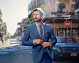 Man in a blue suit, perfect cocktail attire, stands on a street with a vintage car.