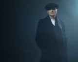 Młody mężczyzna w czapce i płaszczu, z charakterystyczną fryzurą w stylu Peaky Blinders, stoi w zadymionym mroku.