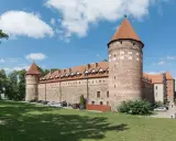 Zamek w Bytowie, potężna ceglana twierdza z charakterystycznymi wieżami i czerwonymi dachami, otoczony zielonym trawnikiem i drzewami.