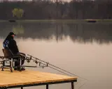 Wędkarz siedzi na wygodnym siedzisku wędkarskim na drewnianym pomoście, czekając na branie.