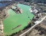 Widok z lotu ptaka na turkusowe jezioro zajączkowskie, otoczone piaszczystymi skarpami i zielenią. Widać sprzęt wydobywczy i łódź.
