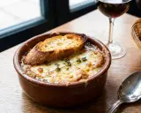Un cuenco de sopa de cebolla gratinada con pan tostado y queso derretido, adornado con tomillo. Una copa de vino tinto y una cuchara completan la escena.