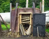 Wypalanie węgla drzewnego w piecu. Drewno ułożone w otwartym palenisku, obok czarne drzwi i inne metalowe konstrukcje.