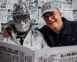 Tomasz Tomaszewski w kapeluszu i płaszczu z gazet, obok uśmiechnięty mężczyzna w czapce z gazet.