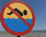 Znak zakazu kąpieli na plaży. Zamknięte kąpieliska nad morzem, mimo pięknej pogody i widocznej w oddali osoby w wodzie.
