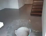 Pavimento continuo de hormigón gris recién aplicado, con una escalera de madera al fondo y un cubo con material.