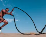Mann mit Kopfhörern macht ein intensives HIIT-Training mit Seilen im Sand unter blauem Himmel.