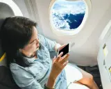 Kobieta siedzi przy oknie samolotu i patrzy na telefon.