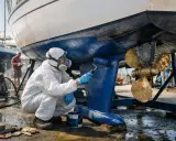 Un homme en combinaison blanche et masque peint la coque bleue d'un bateau. Le calle bateau est en cours de rénovation.