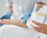 Pielęgniarka w niebieskich rękawiczkach zakłada wenflon do ręki pacjenta, przygotowując kroplówkę.